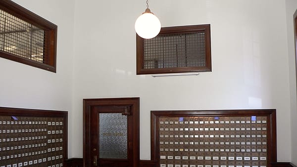 An empty wooden hearing room with a raised bench, microphones on stands and rows of vacant public seating.