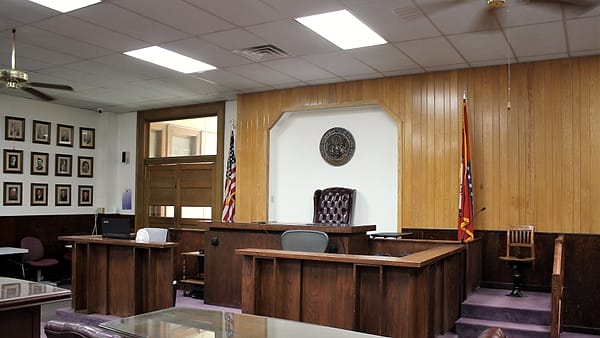 An empty witness chair beside a wooden rail in a courtroom, with a microphone angled toward the seat