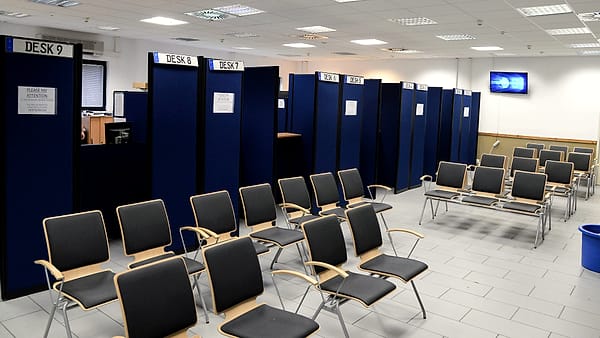 A line of applicants waiting on plastic chairs in a plain corridor, clipboards resting on their knees.
