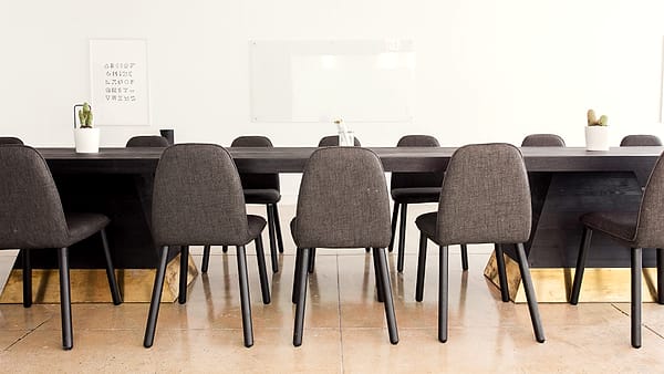 Three empty upholstered chairs set behind a long table in a plain conference room with closed blinds