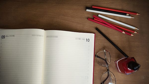 A blank paper wall planner with ruled monthly columns pinned above a desk, its lower corner slightly curled.