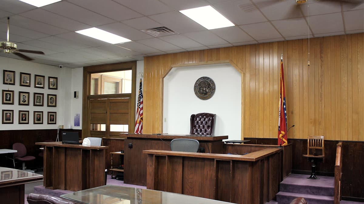 An empty witness chair beside a wooden rail in a courtroom, with a microphone angled toward the seat