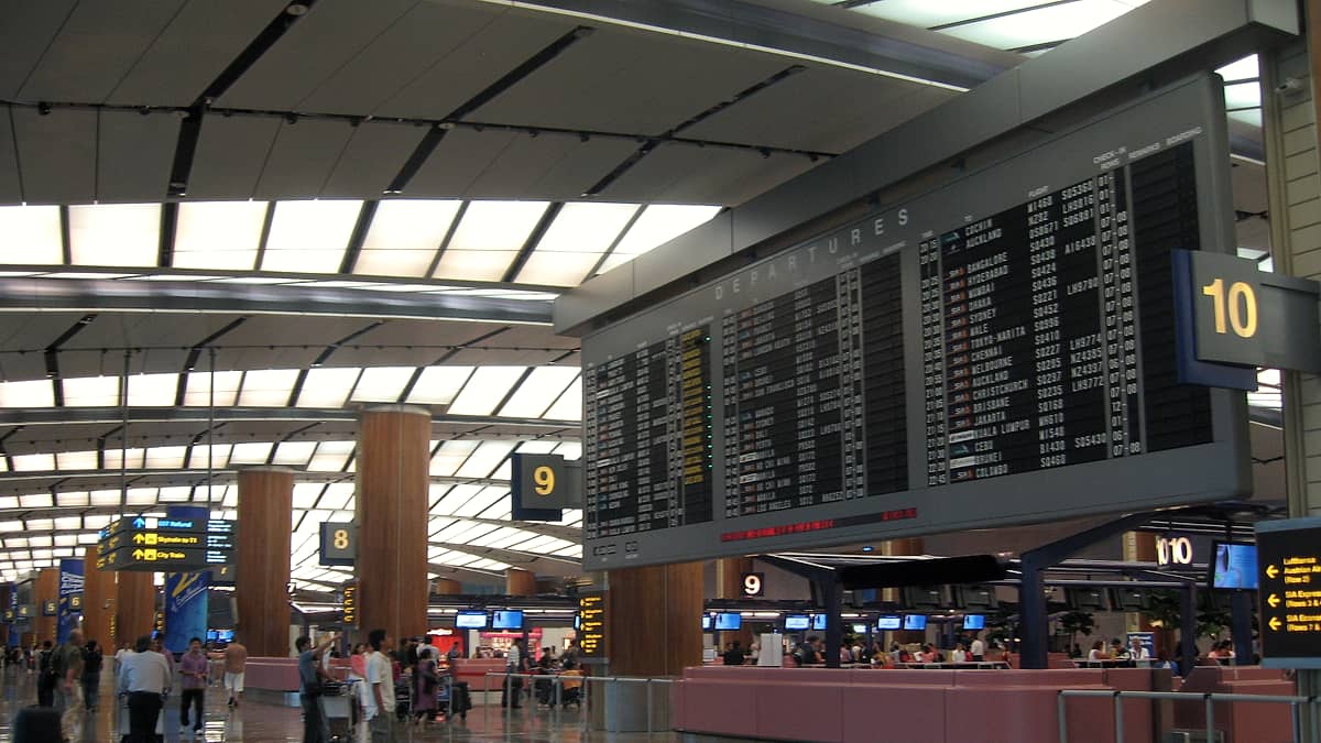 A departures board in an airport terminal listing several cities, with travelers passing beneath it in motion.